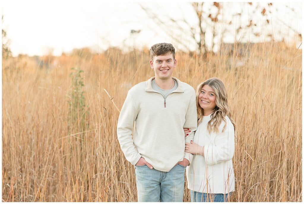 couple standing in field of tall dried grasses and smiling at camera for this Overlook Park Fall Engagement Session