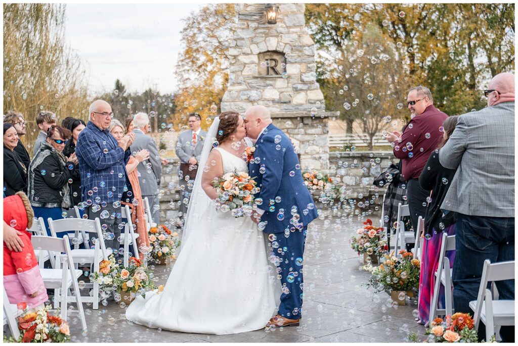 couple kissing in bubbles by guests as they leave ceremony for this Barn at Silverstone Fall Wedding