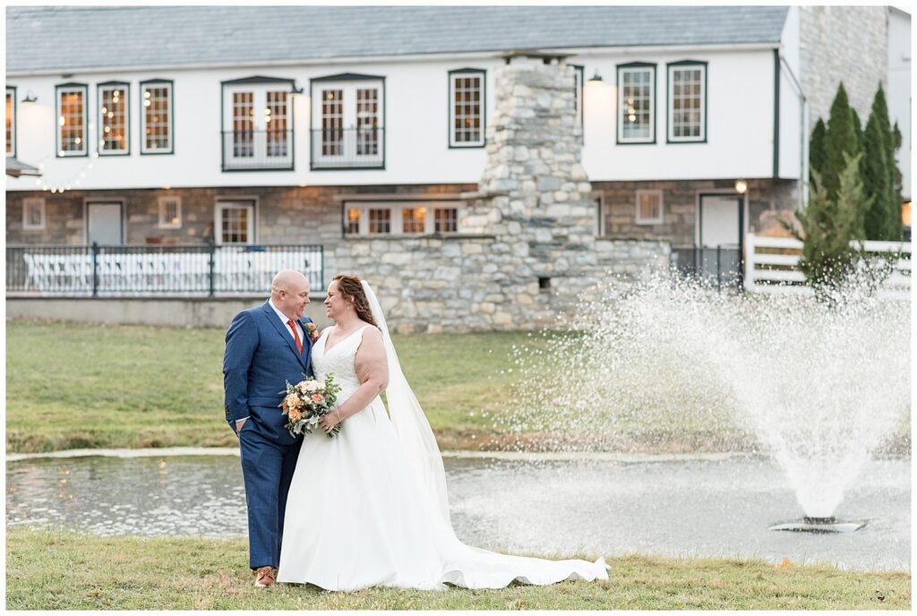 couple standing by pond with barn in background for this Barn at Silverstone Fall Wedding