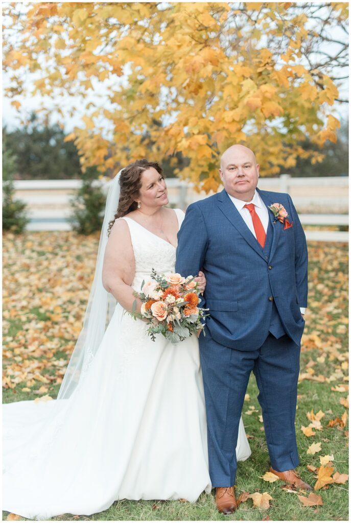 bride holding grooms right arm and smiling at him by colorful tree for this Barn at Silverstone Fall Wedding
