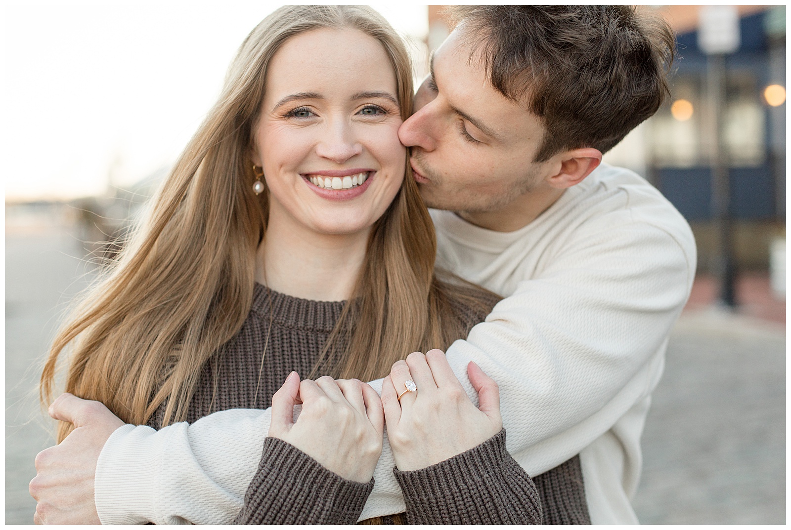 guy hugging girl from behind and kissing her left cheek as she smiles at camera for this Fells Point Engagement Session