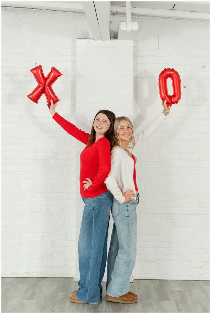 two senior girls extending an X and an O balloon high above them at the white room for this Valentines Day BFF Shoot