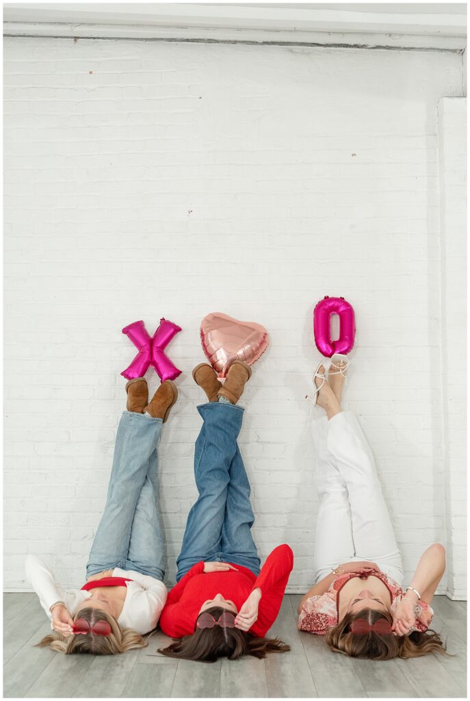 senior girls laying on the floor with their legs extended up white wall and balloons propped on their feet for this Valentines Day BFF Shoot