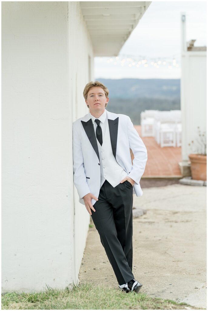 groom leaning against white building with his left hand in his pocket for this Wedding at The Rotunda at Lauxmont Farms