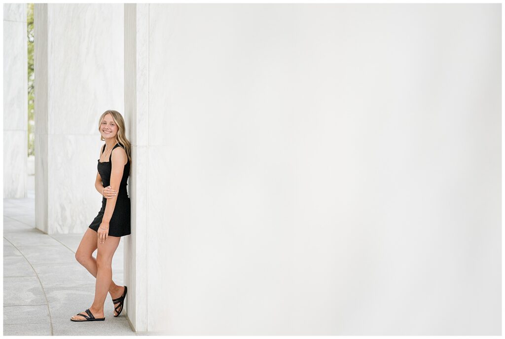 high school senior girl leaning against white column in short black dress