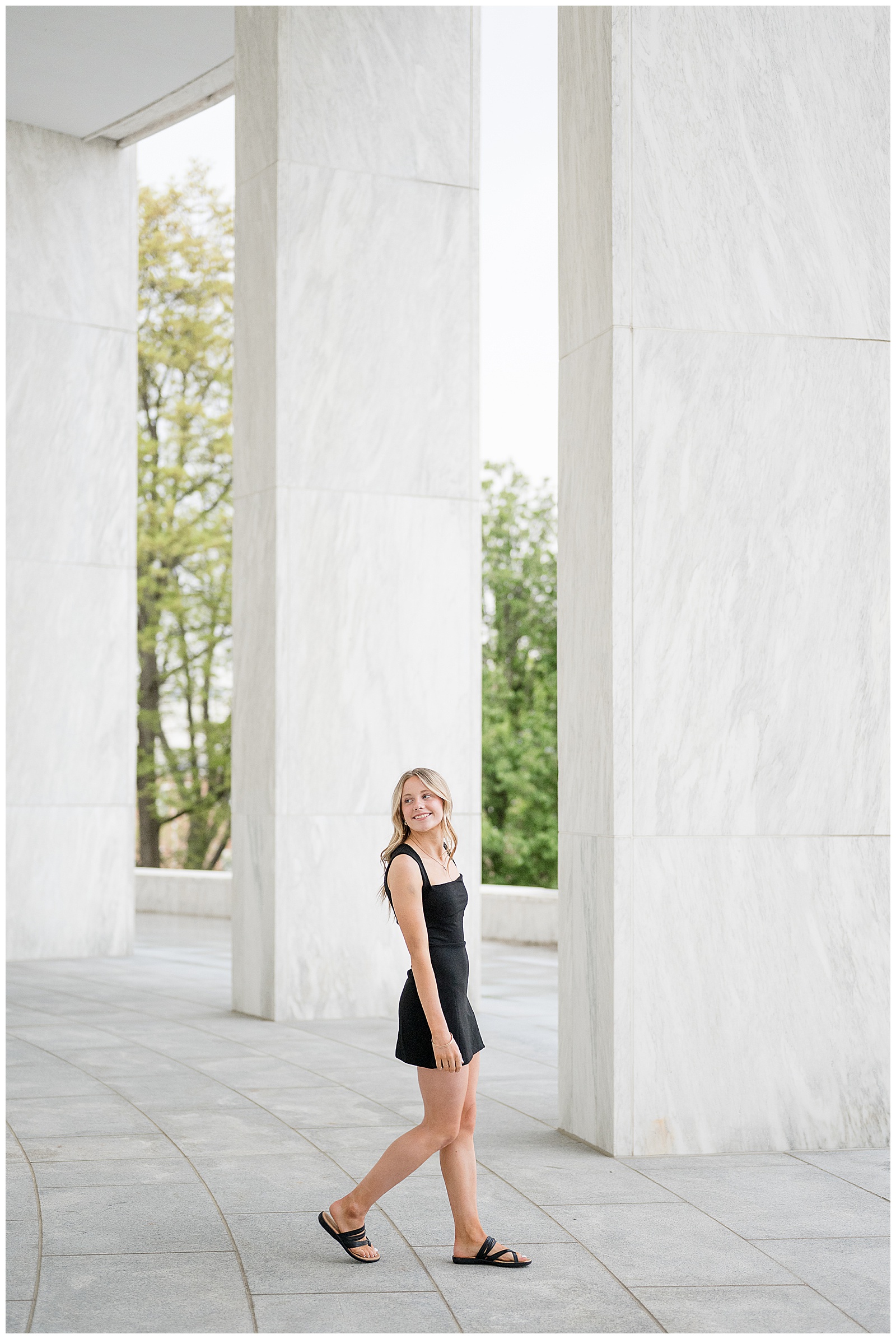 high school senior girl walking past camera smiling over shoulder in short black dress