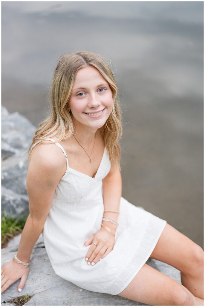high school senior sitting on rock at edge of pond with white dress on