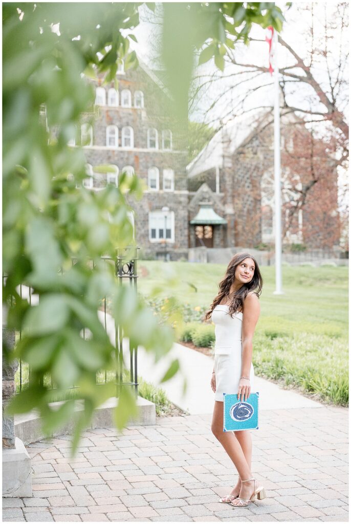 senior looking over shoulder in white dress holding Penn State decorated graduation cap
