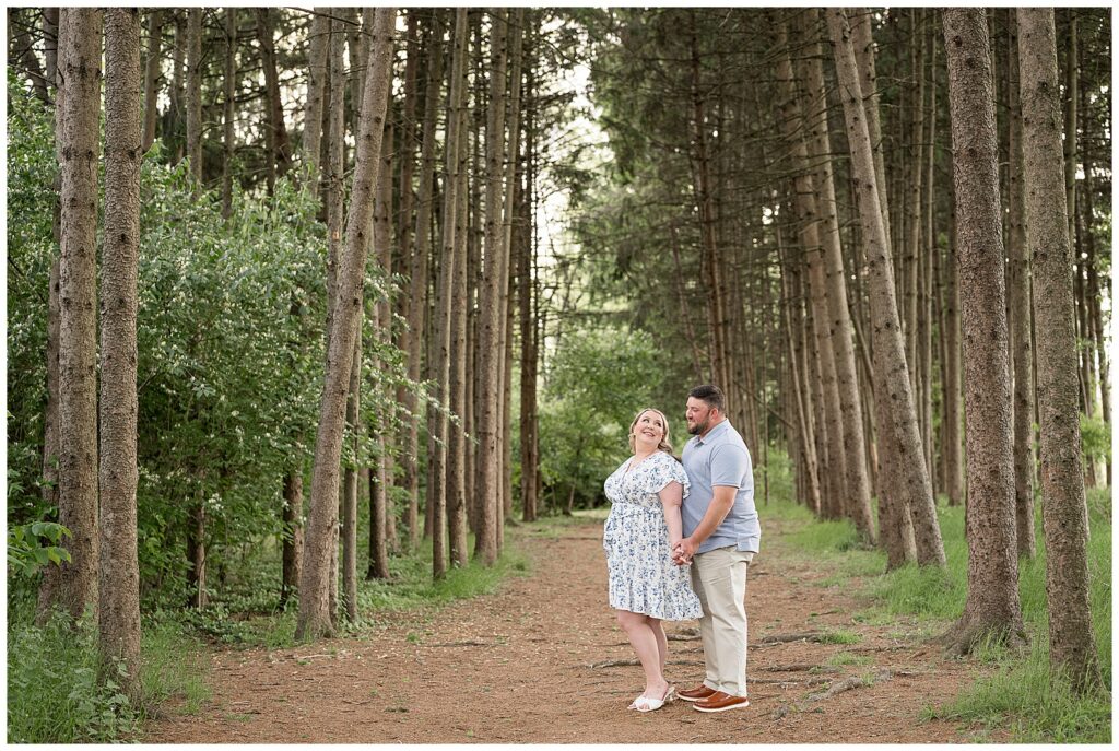 couple standing between rows of pine tree groves holding hands looking at each other