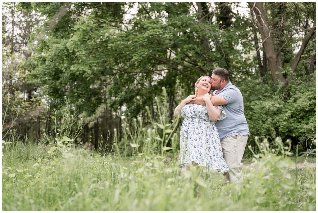guy standing behind girl with his arm wrapped around her and giving kis on cheek