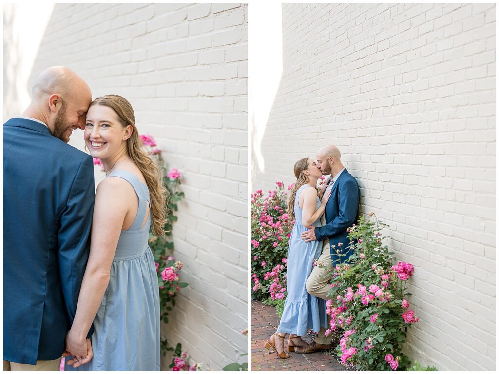 Engaged couple posing in front of a white brick wall with blooming pink climbing roses in downtown Lancaster Pennsylvania, romantic engagement portraits