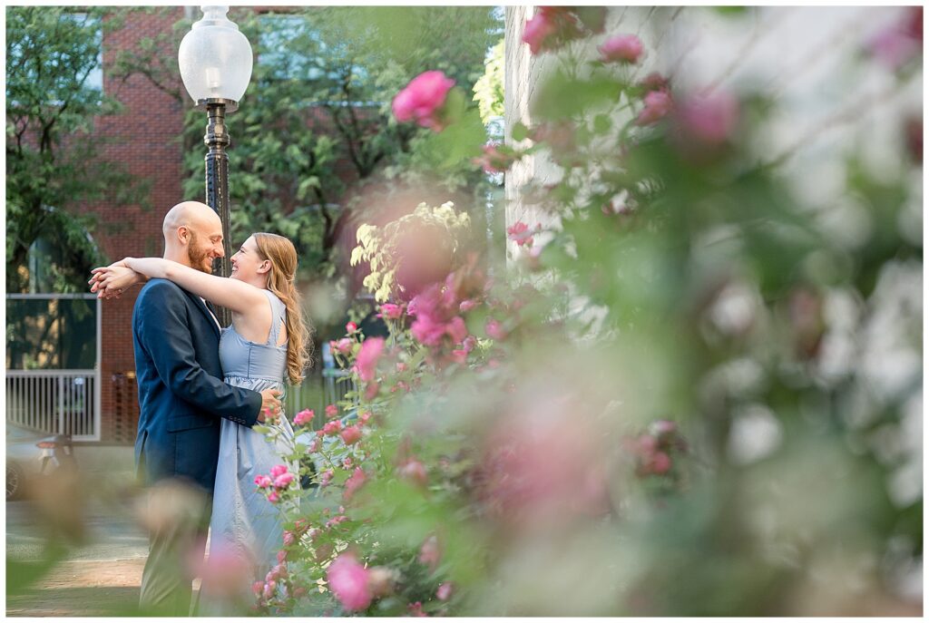 Engaged couple embracing and laughing surrounded by pink roses near an antique street lamp in downtown Lancaster PA engagement session