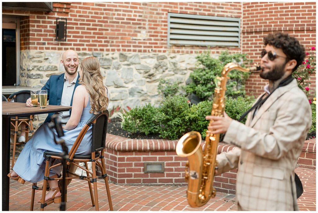 Engaged couple laughing at South County Brewery in Lancaster Pennsylvania while a saxophonist performs on the brick patio, creative engagement photography