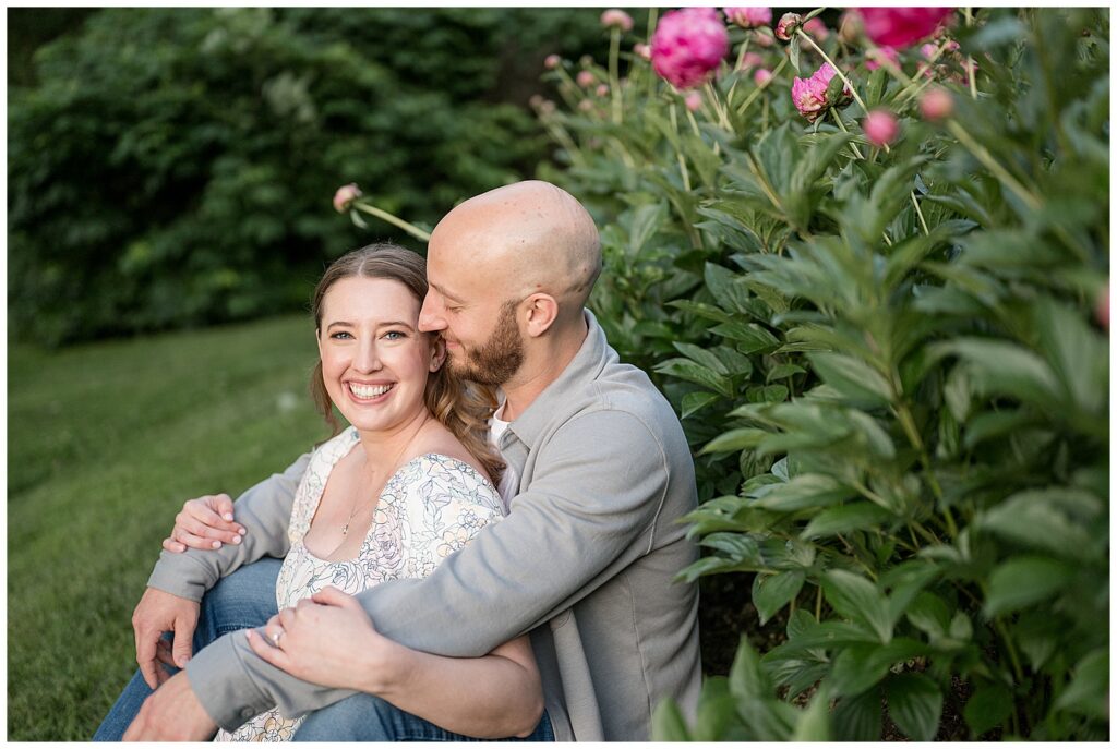 Engaged couple sitting together in front of lush pink peony bushes at Lancaster County Central Park Pennsylvania, romantic summer engagement portraits