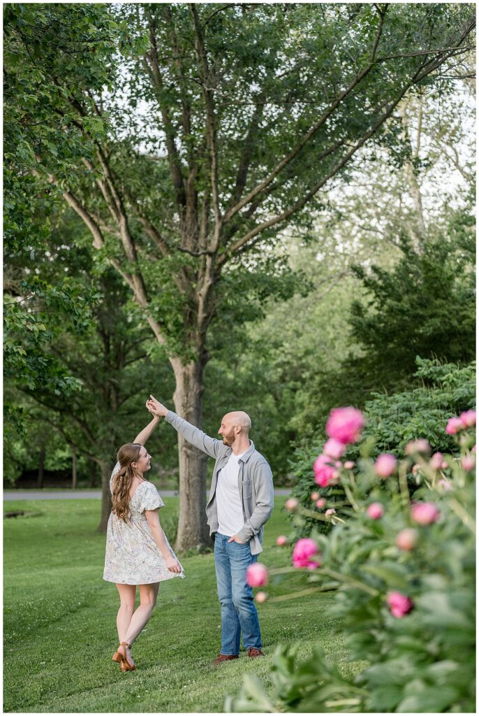 Engaged couple twirling and dancing in an open green field with pink peonies at Lancaster County Central Park PA, playful engagement photography by Heather Marie Photography