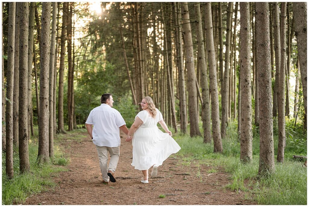 Engaged couple walking hand in hand away from the camera through a sunlit pine tree grove at Overlook Park Lancaster Pennsylvania, candid summer engagement photos