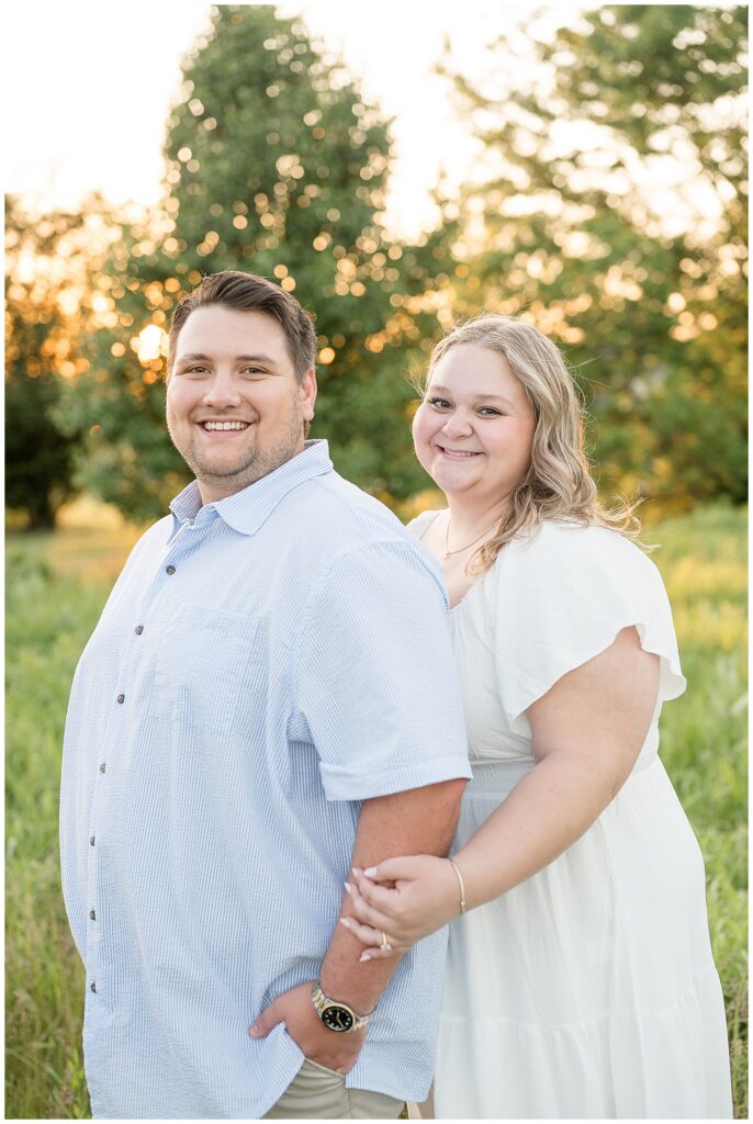Engaged couple smiling directly at the camera in a sun-drenched green field at Overlook Park Lancaster PA, warm summer engagement portraits by Heather Marie Photography