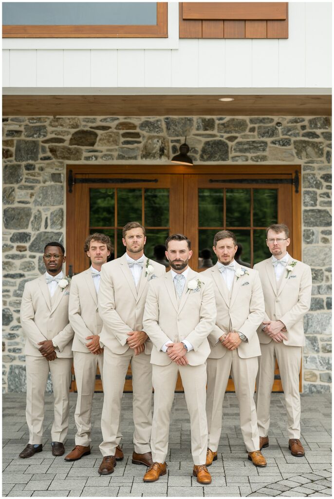 Groomsmen in tan suits with dusty blue ties lined up in a formal portrait in front of the stone barn at Bluestone Estate summer wedding | Heather Marie Photography