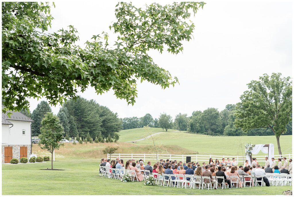 Wide view of the outdoor summer wedding ceremony at Bluestone Estate with rows of white chairs on the green lawn and rolling hills as the backdrop | Heather Marie Photography