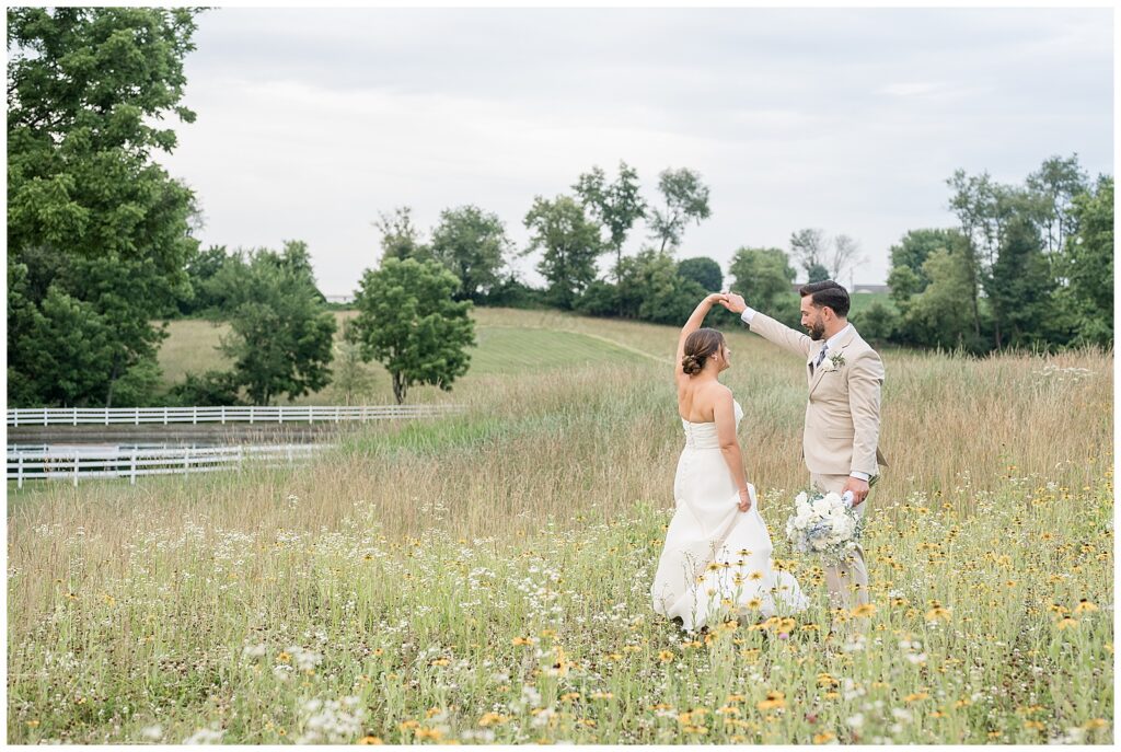 Groom spinning his bride in a wildflower meadow at Bluestone Estate summer wedding, with the white fence and pond glimmering in the background | Heather Marie Photography