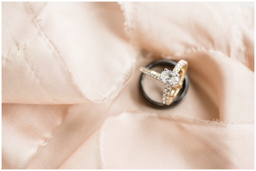 Close-up of a diamond engagement ring with oval center stone and pavé band nested inside a black ceramic groom's wedding band, resting on blush satin fabric, Heather Marie Photography
