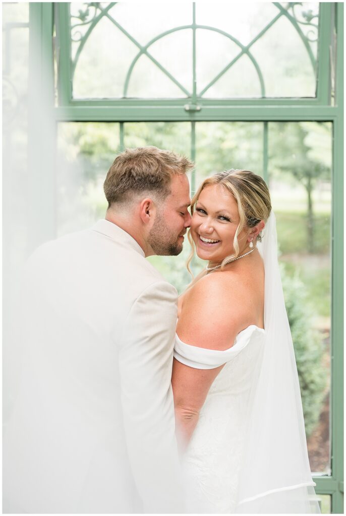 Groom nuzzling bride's cheek as she laughs toward the camera, photographed through the ornate green ironwork windows of the Stone Gables solarium, Heather Marie Photography