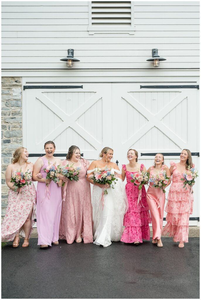 Bride and six bridesmaids in mismatched shades of pink laughing together while holding colorful wildflower bouquets in front of white barn doors at Stone Gables Estate, Heather Marie Photography