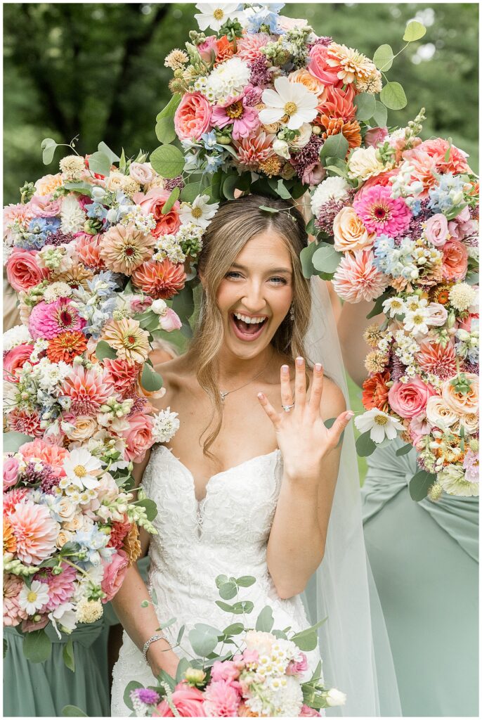 Bride in strapless lace gown laughing and showing wedding ring surrounded by colorful bridesmaid bouquets at Riverdale Manor