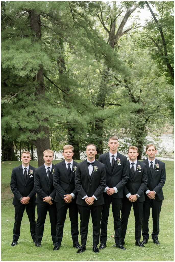 Groomsmen in black suits standing in front of pine trees along the river at Riverdale Manor