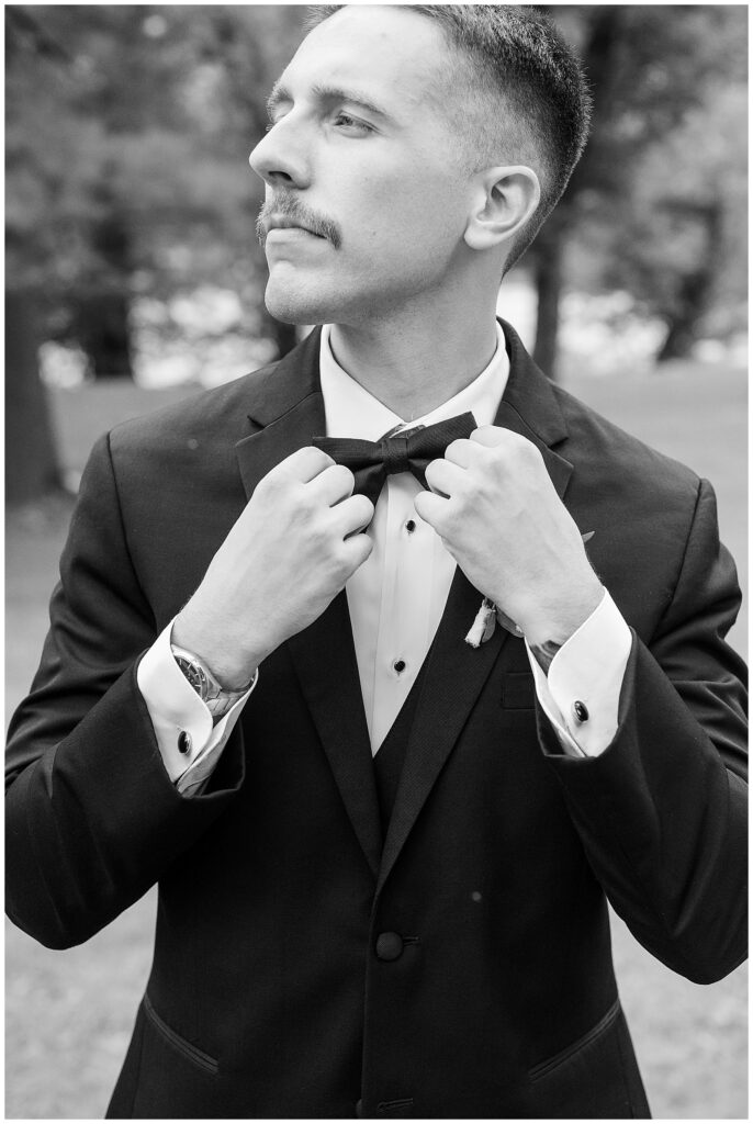 Black and white close up portrait of groom adjusting bow tie in black tuxedo at Riverdale Manor