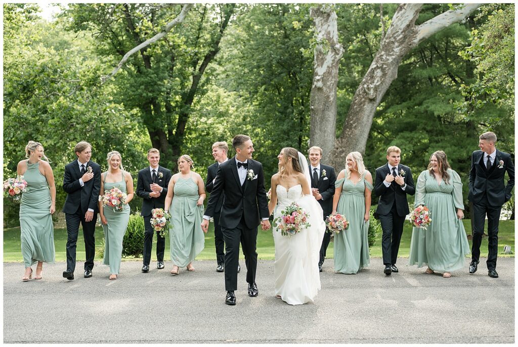 Full wedding party walking portrait with sage green bridesmaids and black tuxedo groomsmen at Riverdale Manor