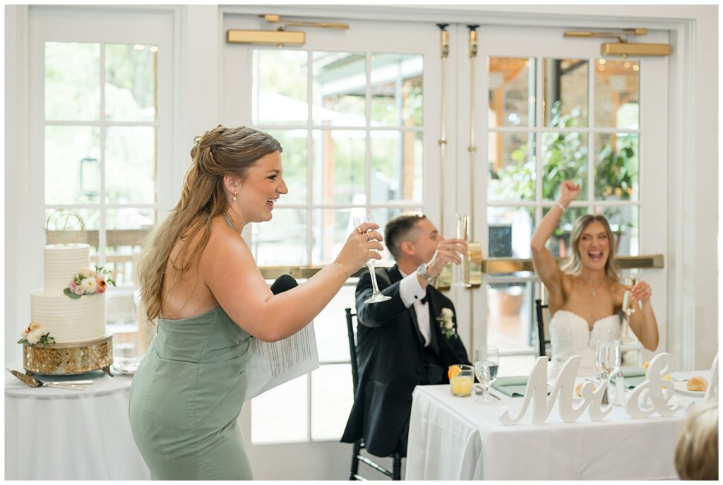 Maid of honor giving toast while bride cheers at Riverdale Manor summer wedding reception