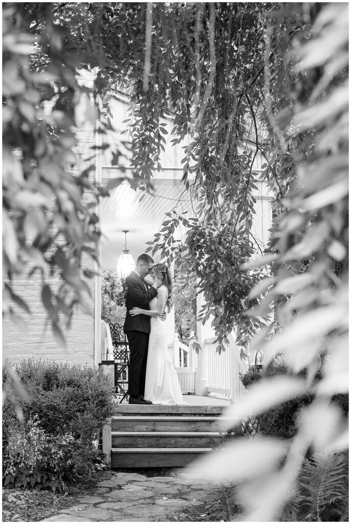 Black and white photo of bride and groom sharing intimate moment on steps framed by weeping willow at Riverdale Manor