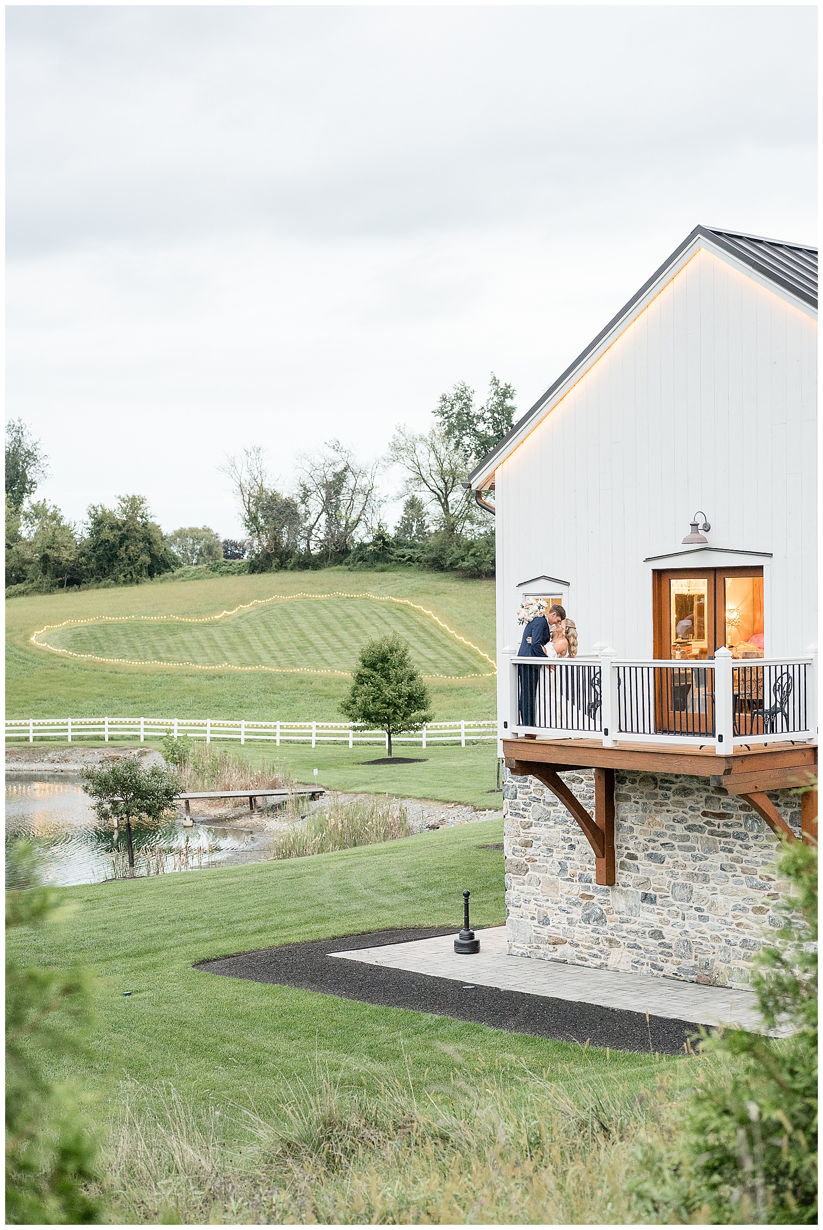 Amanda and Jake kissing on the balcony of the white barn, with a lit heart-shaped lawn, a white fence, and the pond behind them at Bluestone Estate