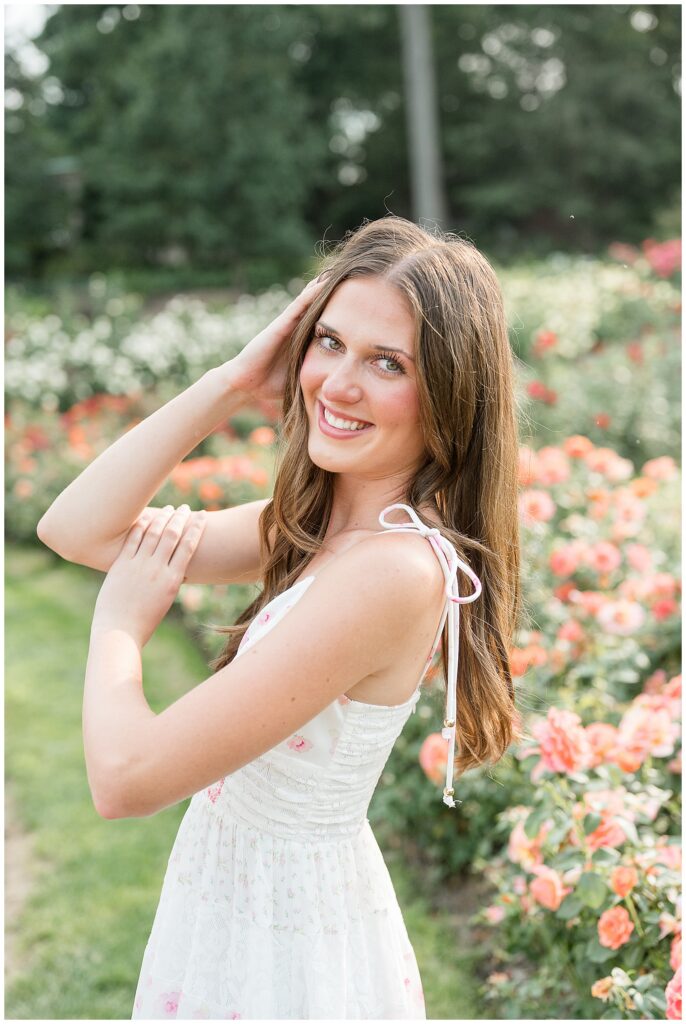 Close-up senior portrait of a girl with green eyes in a white floral dress surrounded by coral roses at Hershey Gardens