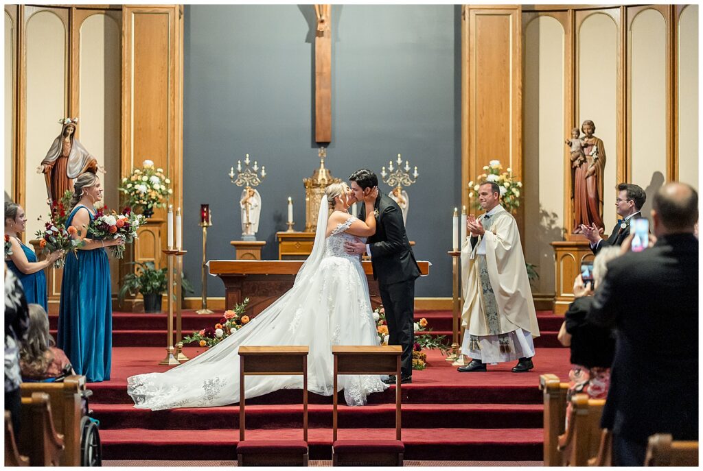 Angela and Ryan sharing their first kiss at the altar of Saint Ignatius church as the priest and bridal party applaud