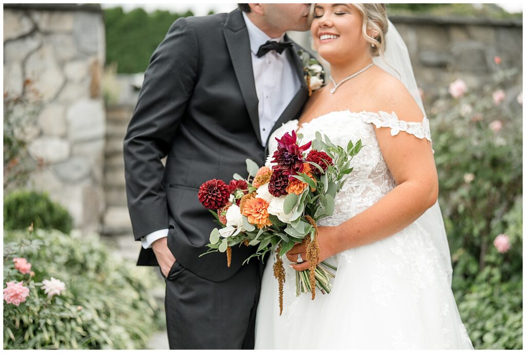 Ryan kissing Angela's temple as she smiles with her dahlia and amaranth bouquet in the rose garden