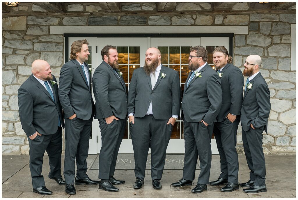 Chris laughing with his groomsmen as they stand with hands in their pockets in front of the stone barn doors